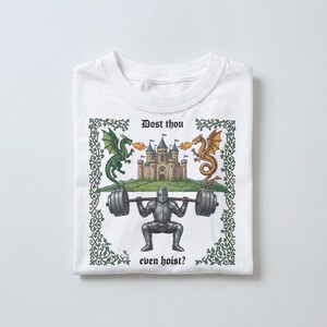 Pu&ograve; includere: T-shirt bianca con grafica a tema medievale. Il disegno presenta un cavaliere che solleva un bilanciere, un castello e due draghi. Il testo recita "Dost thou even hoist?" in un bordo decorativo.