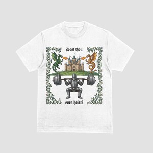Pu&ograve; includere: T-shirt bianca con grafica a tema medievale. Il design include un cavaliere che solleva pesi, un castello e due draghi. Il testo recita "Dost thou even hoist?" circondato da un bordo decorativo.