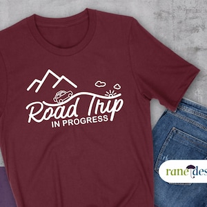 May include: A maroon t-shirt with a white graphic design of a car driving over a mountain road with the text "Road Trip in Progress".