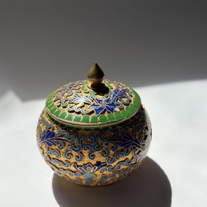 May include: A round, lidded container featuring intricate gold metalwork with blue and green enamel floral patterns. The lid has a pointed finial and a perforated design, while the body displays a continuous, swirling filigree motif.