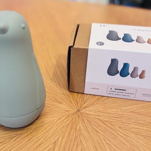 Puede incluir: Un juguete con forma de pingüino azul claro y una caja de cartón con una etiqueta blanca. La etiqueta muestra un conjunto de juguetes de pingüinos en varios colores. La caja está etiquetada como "The Sunshine Penguin Set".