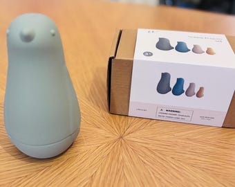 Puede incluir: Un juguete con forma de pingüino azul claro y una caja de cartón con una etiqueta blanca. La etiqueta muestra un conjunto de juguetes de pingüinos en varios colores. La caja está etiquetada como "The Sunshine Penguin Set".