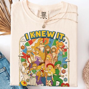 May include: A cream-colored t-shirt featuring a graphic print with the text "I KNEW IT," depicting a group of illustrated female characters in various outfits against a colorful rainbow sunburst background with clouds and flowers.