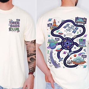 May include: Cream-colored t-shirt with a front pocket design that reads "Tired Dads Club" and a back graphic of a space-themed map with planets, rockets, and aliens. The shirt is a casual wear item.