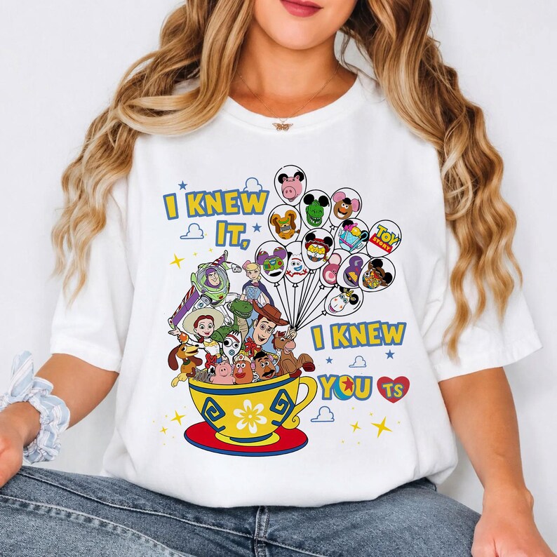 May include: A white t-shirt featuring a colorful graphic of Toy Story characters inside a yellow teacup. The design includes balloons with character faces, yellow text that reads I KNEW IT, I KNEW YOU TS, and small blue stars.
