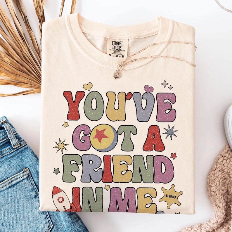 May include: A cream-coloured t-shirt featuring the text "YOU'VE GOT A FRIEND IN ME" in large, multi-coloured, distressed-style letters. The design includes small graphic elements such as a rocket, a sheriff star, hearts, and various star shapes.