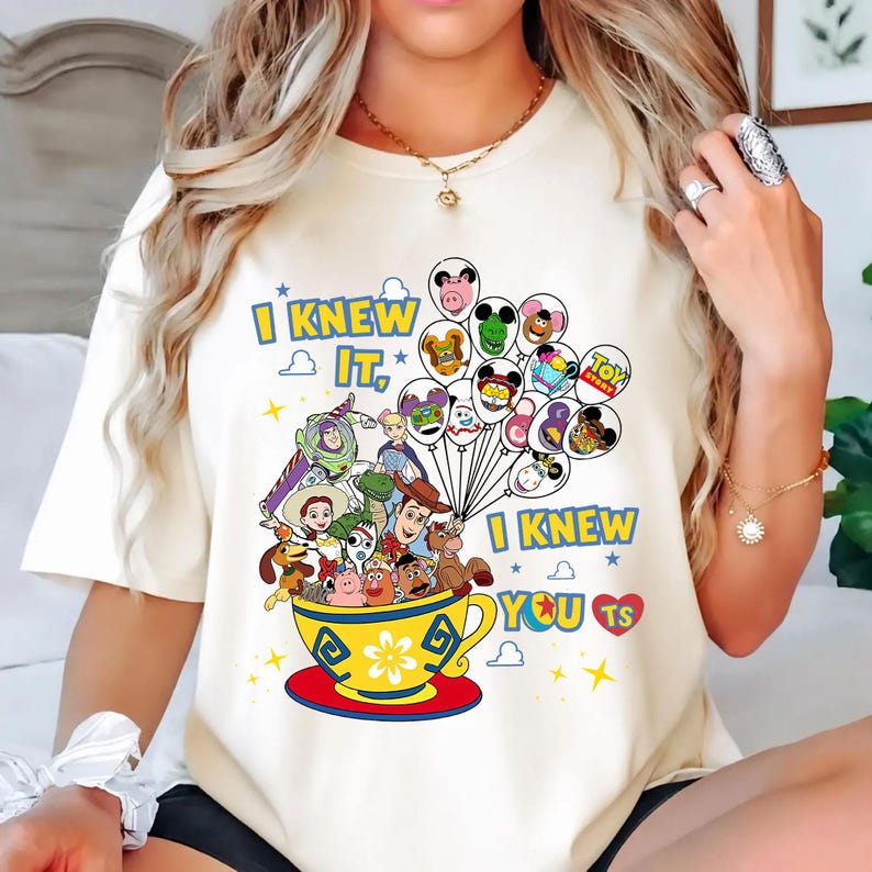 May include: A cream-colored t-shirt featuring a graphic of Toy Story characters inside a yellow teacup. The text reads I KNEW IT, I KNEW YOU TS. A cluster of balloons with character faces floats above the cup, surrounded by small stars and a red heart.