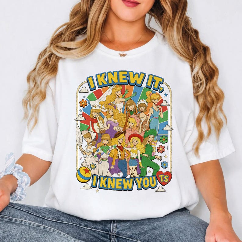 May include: A white t-shirt featuring a colorful graphic print of various animated characters in a retro style. The text "I KNEW IT, I KNEW YOU TS" is printed in bold blue letters above and below the illustration, which includes flowers, clouds, and a ball.