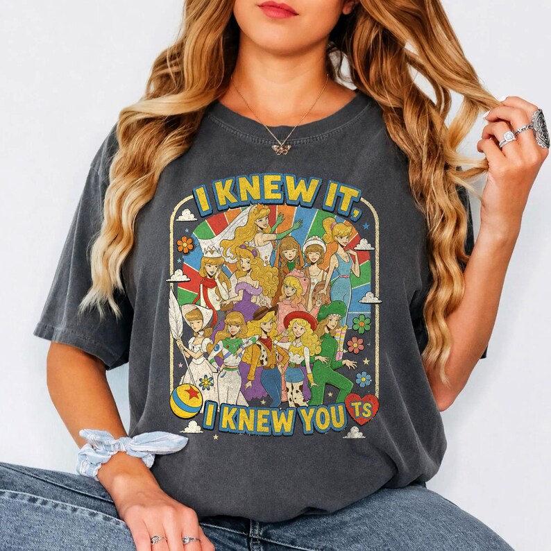 May include: A charcoal gray short-sleeve t-shirt featuring a colorful graphic print of various animated female characters. The text "I KNEW IT, I KNEW YOU TS" is printed in yellow above and below the illustration, which includes flowers and a ball.