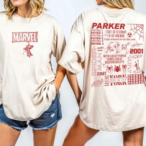 May include: Beige t-shirt featuring a red Marvel logo and Spider-Man graphic on the front. The back displays a Spider-Man design with text including "Parker", "Spider-Man", "2001", and "New York".