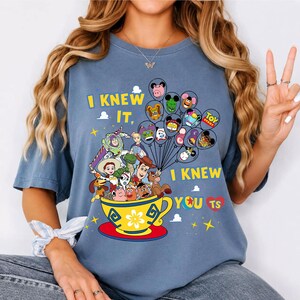 May include: A blue t-shirt featuring a graphic of Toy Story characters inside a yellow teacup with the text I KNEW IT, I KNEW YOU TS. The design includes a cluster of balloons shaped like various characters and stars surrounding the central illustration.