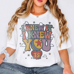 Pu&ograve; includere: Una t-shirt bianca a maniche corte con una stampa grafica multicolore con il testo I KNEW IT I KNEW YOU. Il design include una palla gialla e blu con una stella rossa, una forma a cuore con le lettere TS e varie piccole stelle e nuvole.