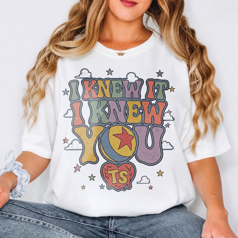 Pu&ograve; includere: Una t-shirt bianca a maniche corte con una stampa grafica multicolore con il testo I KNEW IT I KNEW YOU. Il design include una palla gialla e blu con una stella rossa, una forma a cuore con le lettere TS e varie piccole stelle e nuvole.