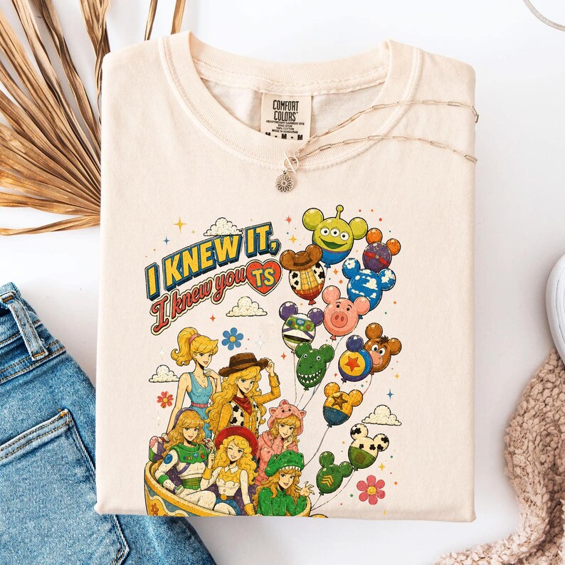 May include: A cream-colored t-shirt featuring a graphic print of various anime-style characters and balloons shaped like cartoon heads. The text reads I KNEW IT, I knew you TS. The shirt has a Comfort Colors brand label at the inner neckline.