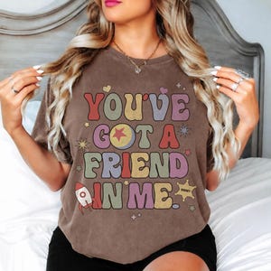 May include: A brown short-sleeve t-shirt featuring the text "YOU'VE GOT A FRIEND IN ME." The graphic includes a rocket, a sheriff star, and various stars in muted colours like red, yellow, green, and purple, arranged in a retro-style font layout.
