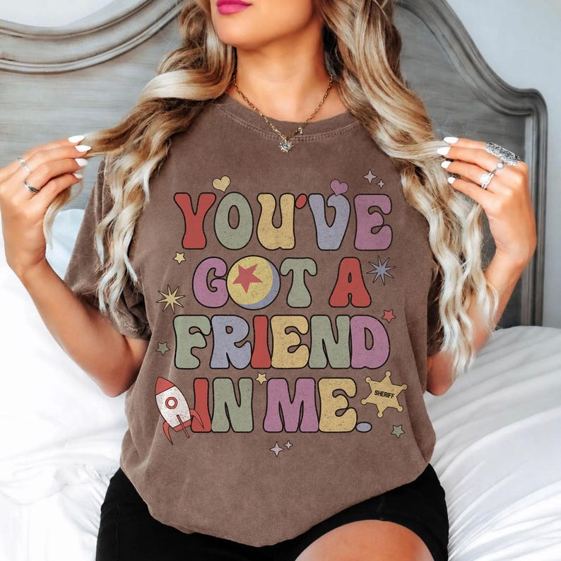 May include: A brown short-sleeve t-shirt featuring the text "YOU'VE GOT A FRIEND IN ME." The graphic includes a rocket, a sheriff star, and various stars in muted colours like red, yellow, green, and purple, arranged in a retro-style font layout.