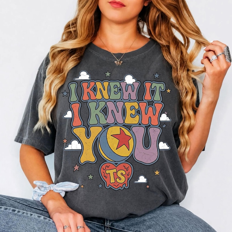 Pu&ograve; includere: Una t-shirt grigio antracite a maniche corte con il testo I KNEW IT I KNEW YOU TS in un carattere retro colorato. Il design include una palla gialla con una stella rossa, un cuore rosso e piccole nuvole e stelle bianche attorno alla scritta.