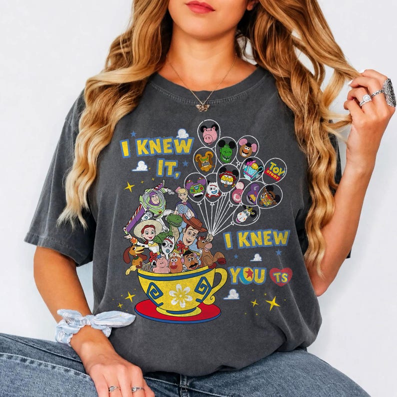 May include: A charcoal gray short-sleeve t-shirt featuring a graphic of Toy Story characters in a yellow teacup with balloons. The text reads I KNEW IT, I KNEW YOU TS. The design includes various colorful animated characters and small yellow stars.