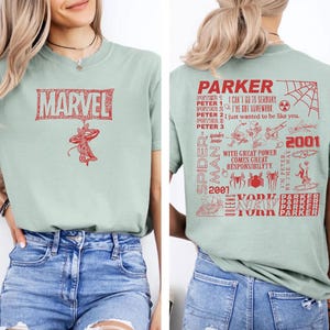 May include: A light green t-shirt featuring a red Marvel logo and a Spider-Man graphic. The back of the shirt has a Spider-Man design with text that reads "Parker" and "2001".