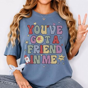 May include: A blue short-sleeve t-shirt featuring the text "YOU'VE GOT A FRIEND IN ME" in a multi-coloured retro font. The graphic includes a small rocket ship, a sheriff star badge, and various stars and hearts in red, yellow, and purple.