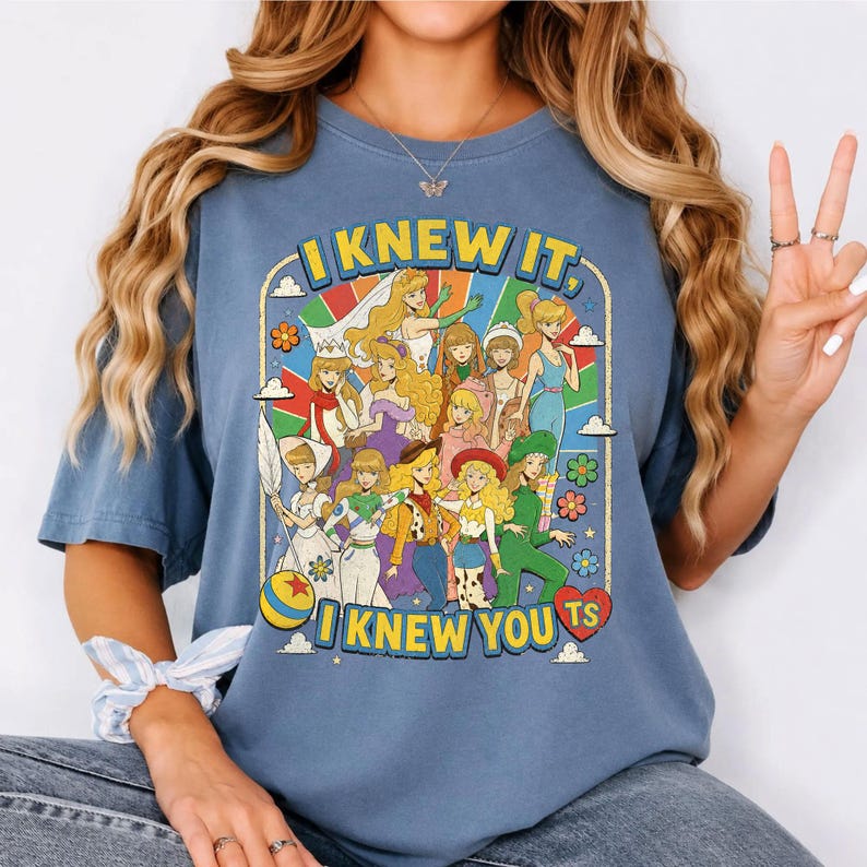 May include: A blue short-sleeve t-shirt featuring a graphic print of various animated female characters in a retro style. The text "I KNEW IT, I KNEW YOU TS" is printed in yellow block letters above and below the colorful illustration.