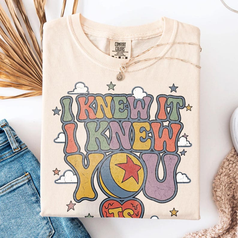 Pu&ograve; includere: Una t-shirt color crema con la scritta I KNEW IT I KNEW YOU TS in un carattere retr&ograve; multicolore. Il design include una palla gialla con una stella rossa, piccole nuvole e varie stelle sparse attorno al testo. Etichetta Comfort Colors presente.