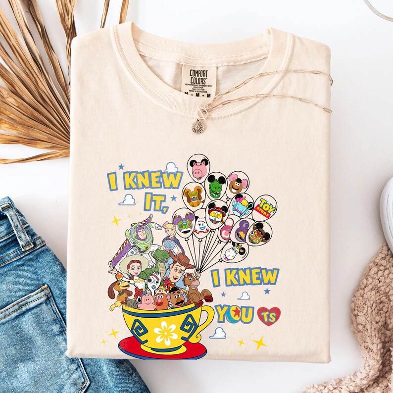 May include: A cream-colored t-shirt featuring a graphic of Toy Story characters inside a yellow teacup, holding a cluster of balloons with character faces. The text reads I KNEW IT, I KNEW YOU TS with a red heart and small yellow stars.