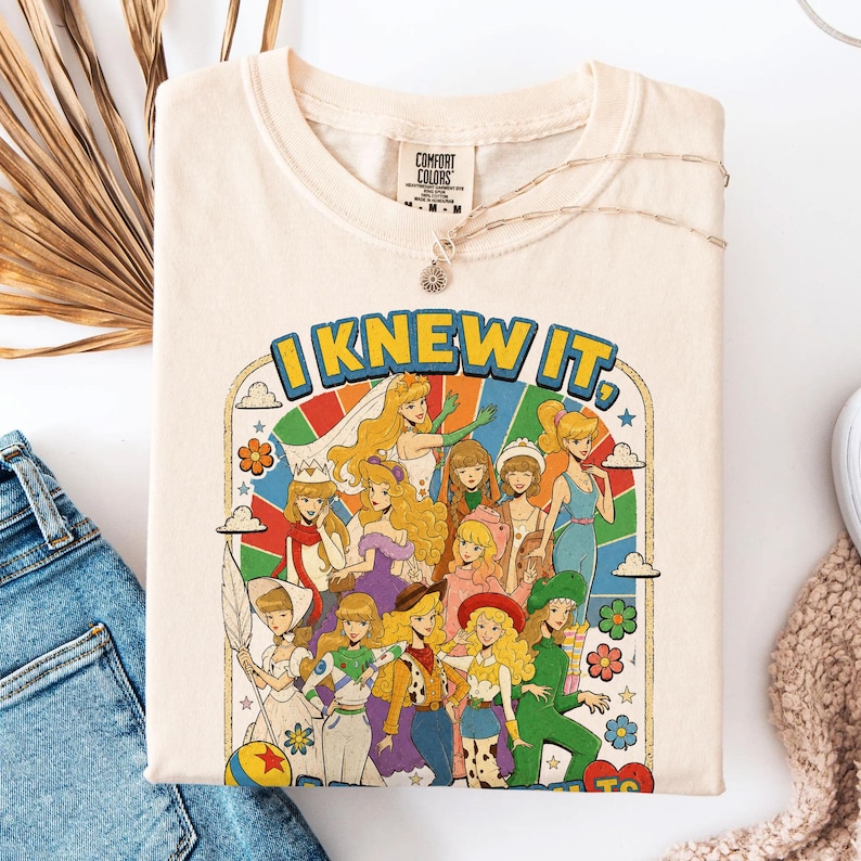 May include: A cream-colored t-shirt featuring a graphic print with the text "I KNEW IT," depicting a group of illustrated female characters in various outfits against a colorful rainbow sunburst background with clouds and flowers.