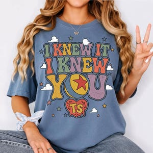 Pu&ograve; includere: Una t-shirt blu a maniche corte con una stampa grafica che riporta il testo I KNEW IT I KNEW YOU. Il design include una palla gialla con una stella rossa, una forma a cuore con le lettere TS e varie piccole nuvole e stelle bianche.