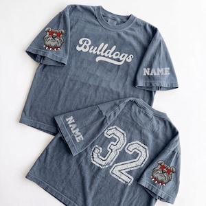 May include: Two blue-grey t-shirts featuring the word Bulldogs in white script on the chest. One sleeve displays a bulldog wearing red star-shaped glasses and a spiked collar, while the other sleeve features the word NAME in white block letters.