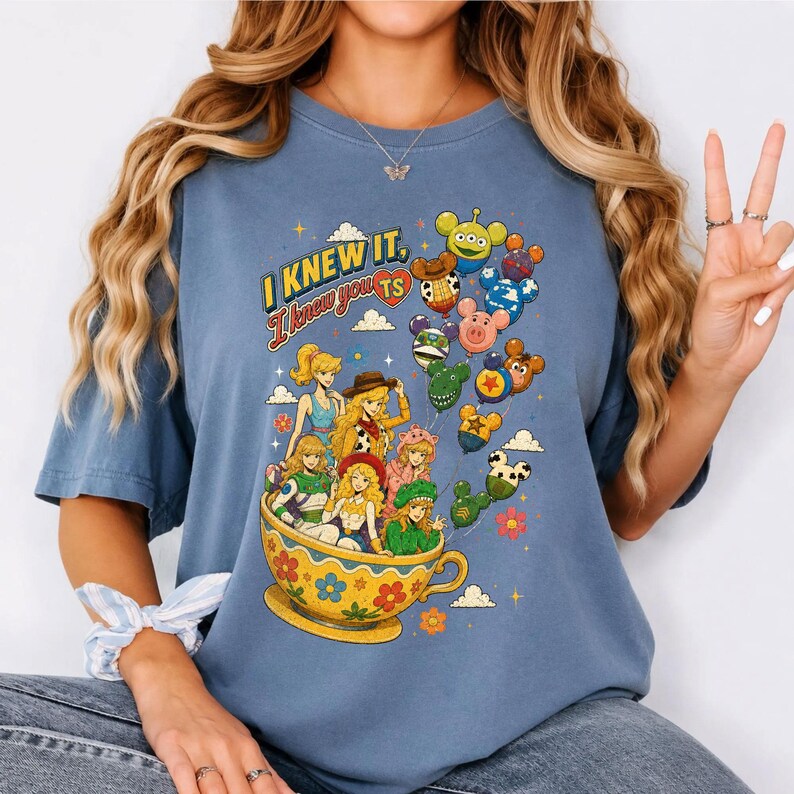 May include: A blue t-shirt featuring a graphic print of various animated characters sitting in a large yellow teacup. The text "I KNEW IT, I knew you" is printed above the characters, accompanied by several colorful balloons shaped like mouse ears.