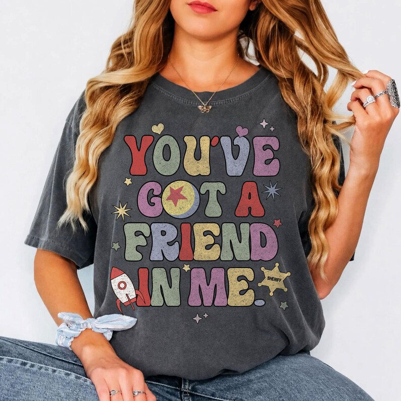 May include: A charcoal grey short-sleeve t-shirt featuring the text "YOU'VE GOT A FRIEND IN ME." in multi-coloured, retro-style bubble letters. The design includes a small rocket ship, a sheriff star badge, and various star and heart icons.