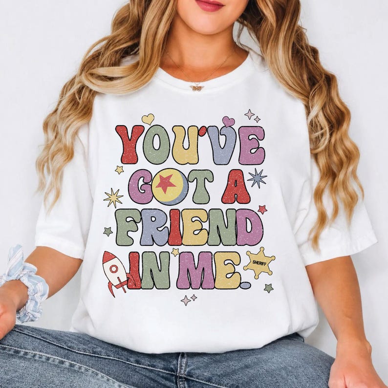 May include: A white short-sleeved t-shirt featuring the text "YOU'VE GOT A FRIEND IN ME." in a multi-coloured, retro-style font. The design includes a yellow ball with a red star, a small red rocket ship, a sheriff's badge, and various stars.