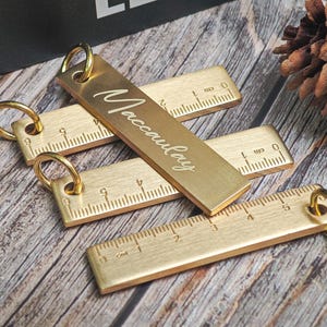 May include: Several brass ruler keyrings with gold rings. One is engraved with the name "Maccauley". The rulers are marked with centimetre measurements and are displayed on a wooden surface.