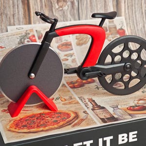 K&ouml;nnte beinhalten: Ein neuartiger Pizzaschneider in Form eines Fahrrads. Der Schneider hat einen roten Rahmen, schwarze R&auml;der und eine kreisf&ouml;rmige Klinge. Er liegt auf einer dekorativen Oberfl&auml;che mit Pizza- und Tomatenillustrationen. Ein lustiges K&uuml;chenwerkzeug.
