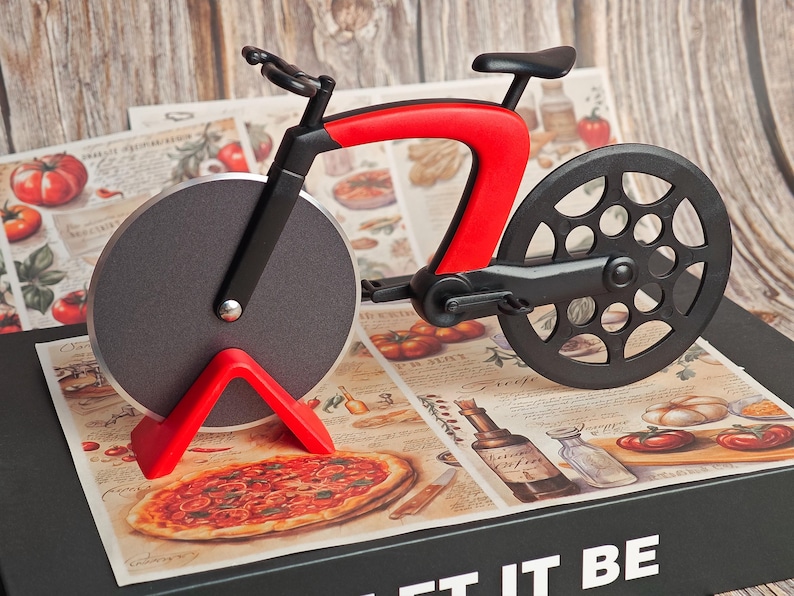 K&ouml;nnte beinhalten: Ein neuartiger Pizzaschneider in Form eines Fahrrads. Der Schneider hat einen roten Rahmen, schwarze R&auml;der und eine kreisf&ouml;rmige Klinge. Er liegt auf einer dekorativen Oberfl&auml;che mit Pizza- und Tomatenillustrationen. Ein lustiges K&uuml;chenwerkzeug.