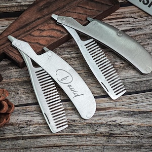Könnte beinhalten: Drei silberfarbene Klappkämme, jeweils mit Kamm und Griff. Auf einem Kamm ist der Name "David" eingraviert. Die Kämme liegen auf einer Holzoberfläche.