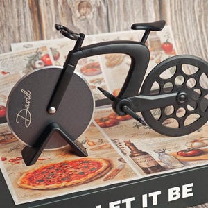 Könnte beinhalten: Ein schwarzer Pizza-Schneider in Form eines Fahrrads. Das Vorderrad ist eine kreisförmige Klinge mit dem Namen "David" in Schreibschrift. Der Pizzaschneider liegt auf einer Box mit Pizza- und Tomaten-Motiv.