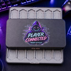 以下が含まれることがあります： 「PLAYER CONNECTED - READY TO PLAY」の文字とフード付きの人物のグラフィックが入った黒いゲームカートリッジケース。ケースは開いており、ゲームカートリッジ用のスロットが見えます。他のゲームアクセサリーが背景にあります。