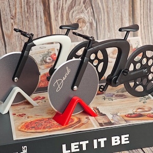 K&ouml;nnte beinhalten: Pizza-Schneider in Form eines Fahrrads. Einer ist schwarz, einer wei&szlig; und einer grau mit dem Namen "David" darauf. Die Pizzaschneider stehen auf einer Box mit der Aufschrift "LET IT BE".