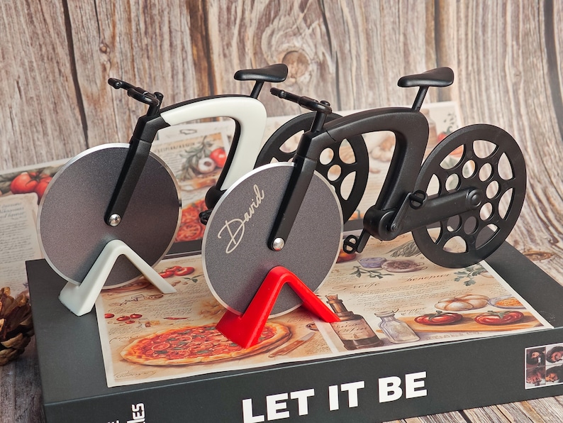 K&ouml;nnte beinhalten: Pizza-Schneider in Form eines Fahrrads. Einer ist schwarz, einer wei&szlig; und einer grau mit dem Namen "David" darauf. Die Pizzaschneider stehen auf einer Box mit der Aufschrift "LET IT BE".