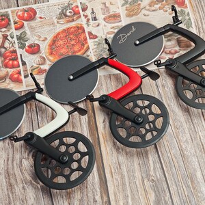K&ouml;nnte beinhalten: Pizza-Schneider-Set in Form von Fahrr&auml;dern. Die Schneider haben schwarze R&auml;der und Klingen, mit roten, wei&szlig;en und schwarzen Rahmen. Auf einem Schneider steht der Name "David" auf der Klinge. Der Hintergrund zeigt eine Pizza-Collage.