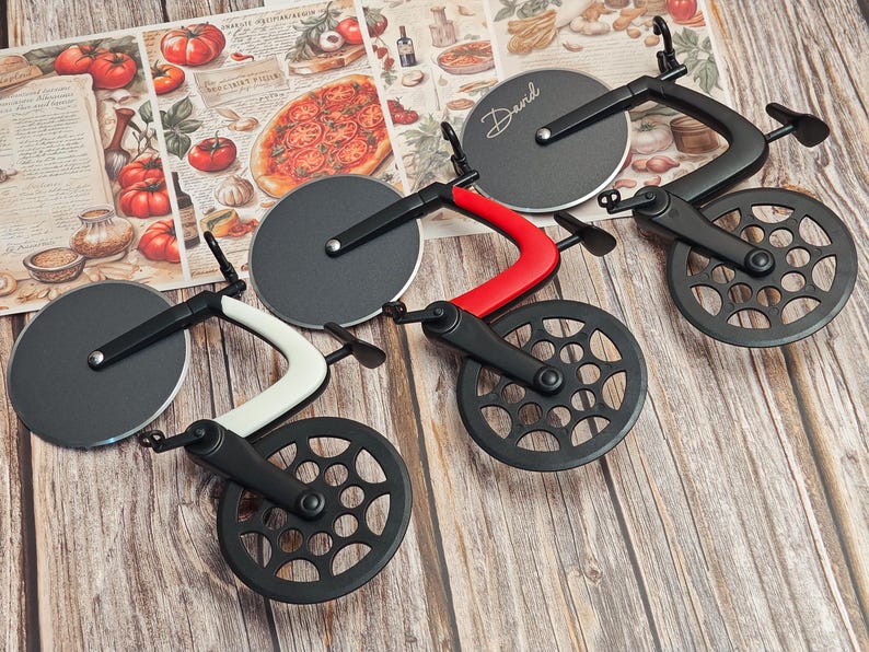 K&ouml;nnte beinhalten: Pizza-Schneider-Set in Form von Fahrr&auml;dern. Die Schneider haben schwarze R&auml;der und Klingen, mit roten, wei&szlig;en und schwarzen Rahmen. Auf einem Schneider steht der Name "David" auf der Klinge. Der Hintergrund zeigt eine Pizza-Collage.
