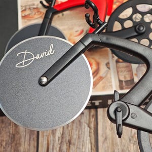 K&ouml;nnte beinhalten: Ein schwarz-roter Pizza-Schneider in Fahrradform mit einer silbernen Klinge. Auf der Klinge steht der Name "David" in wei&szlig;er Schrift. Der Pizzaschneider hat einen schwarzen Rahmen und R&auml;der.