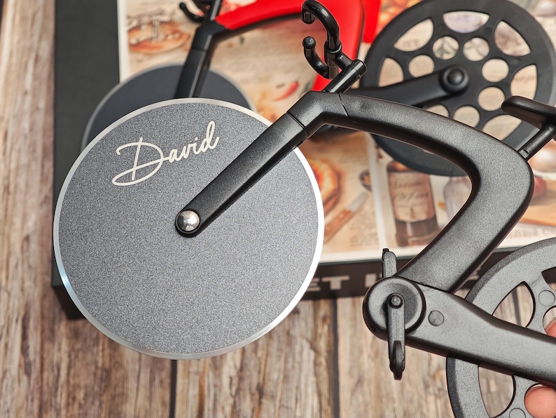 K&ouml;nnte beinhalten: Ein schwarz-roter Pizza-Schneider in Fahrradform mit einer silbernen Klinge. Auf der Klinge steht der Name "David" in wei&szlig;er Schrift. Der Pizzaschneider hat einen schwarzen Rahmen und R&auml;der.