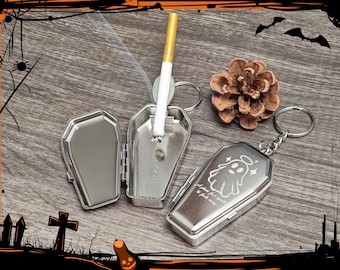 May include: A silver coffin-shaped cigarette case with a lit cigarette, a pinecone, and a matching keychain. The keychain case has a ghost illustration and the text "A place for spirits to find safety."