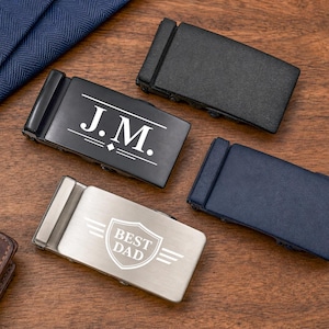 May include: Four rectangular belt buckles in black, navy blue, and brushed silver finishes. One black buckle features the engraved text J.M. with decorative lines, and one silver buckle displays a shield graphic with the text BEST DAD.
