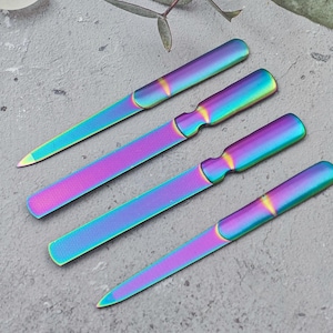 Könnte beinhalten: Vier schillernde Nagelfeilen liegen auf einer grauen Oberfläche. Die Feilen haben eine rechteckige Form mit abgerundeten Enden und einem griffartigen Design. Die Farbe wechselt zwischen Lila, Blau und Grün.