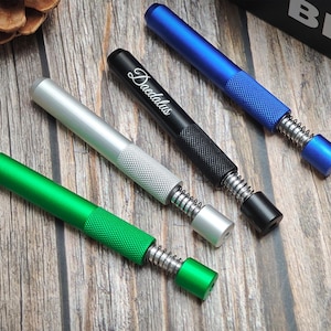 Op de afbeelding: Vier metalen rookaccessoires in groen, zilver, zwart en blauw. De zwarte heeft het woord "Daedalus" in script. Elk heeft een getextureerde grip en een veermechanisme.