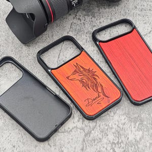 May include: Three phone cases with black frames. One case is black, one has a red wood grain design, and the other has a wood grain design with a wolf engraving and the name "David" below it. A camera lens is in the background.